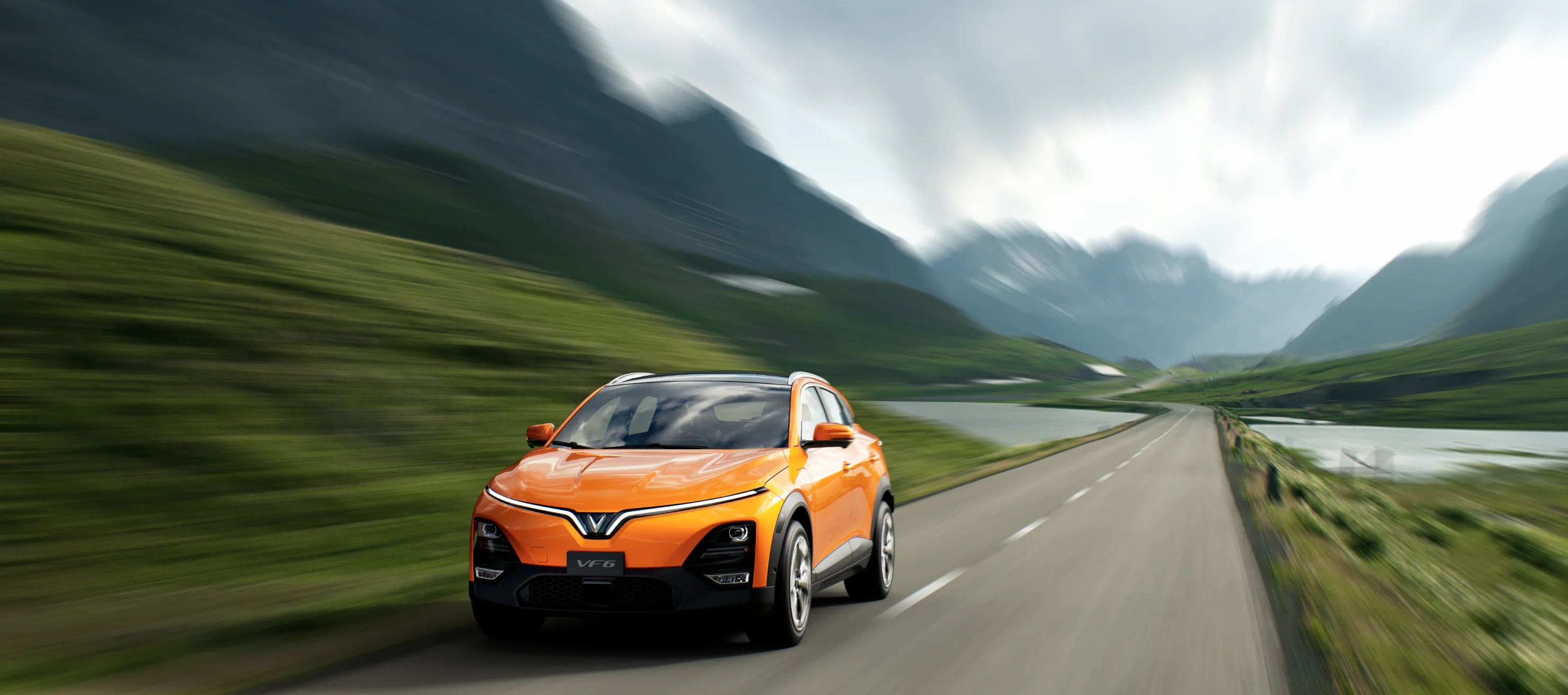 A sunset Orange VF 6 is driving fast down the road near a mountain landscape