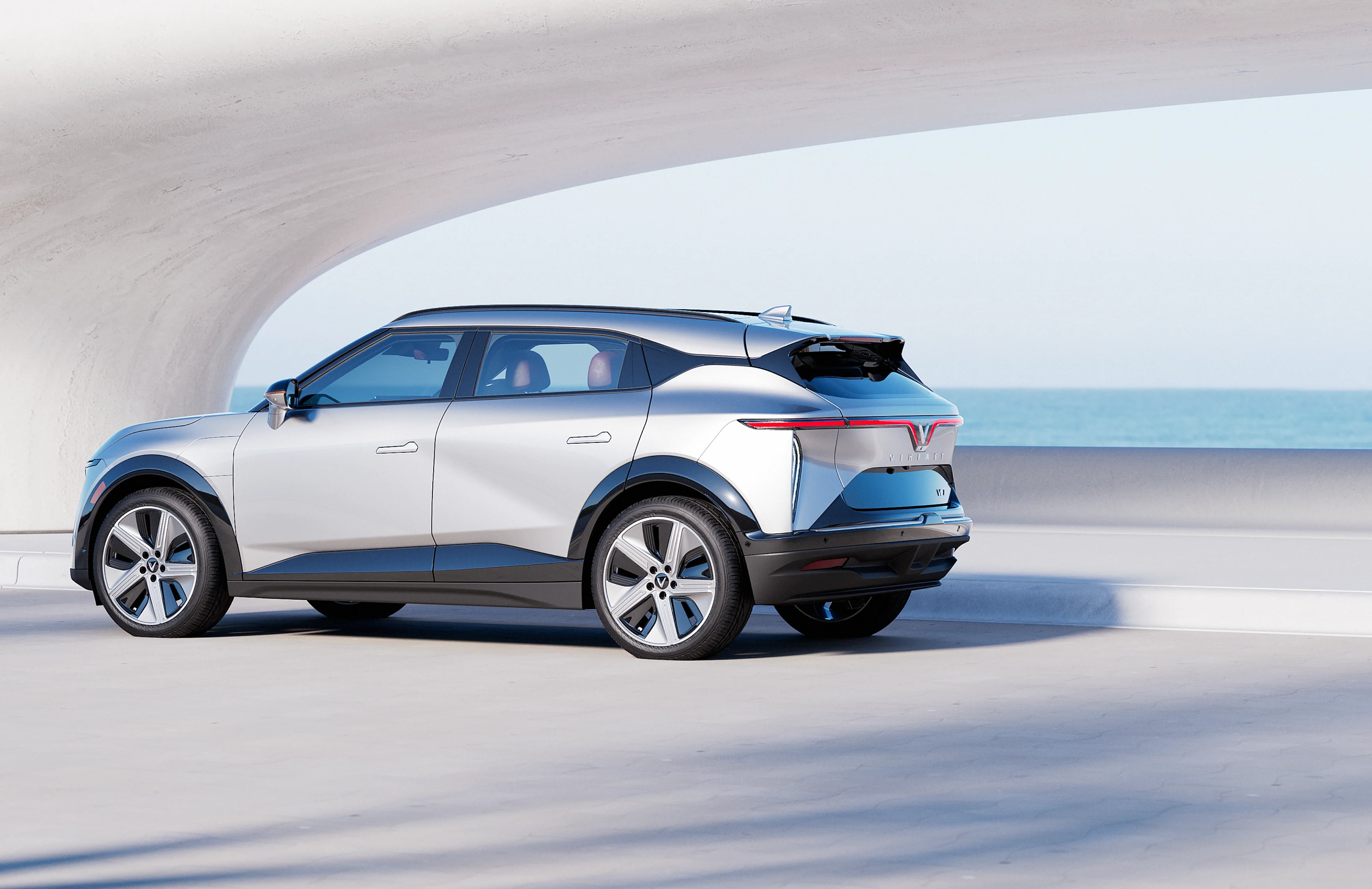 A Desat Silver VinFast VF 7 is parked on the road near the ocean on a sunny day