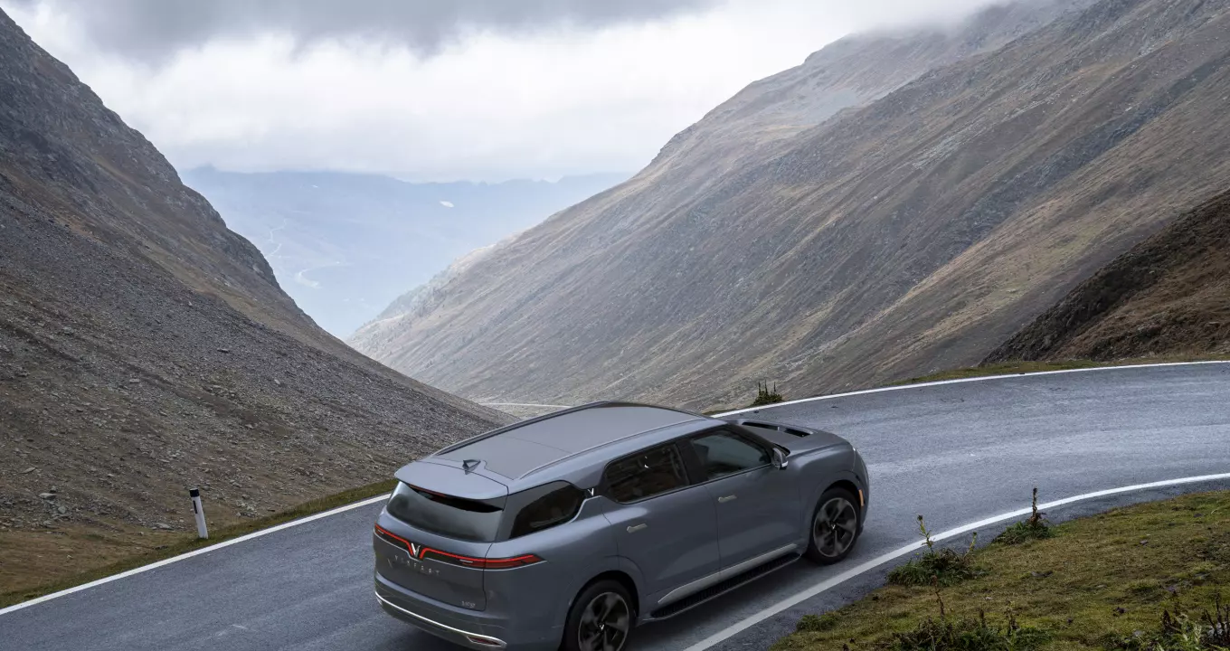 A gray VinFast VF 9 is driving up a curved road in the mountains on a cloudy day